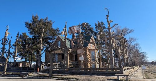 A unique log cabin surrounded by bare trees, adorned with various sculptures and a flag, under a clear blue sky.