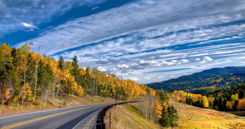A winding road through vibrant autumn trees under a blue sky with wispy clouds and distant mountains.
