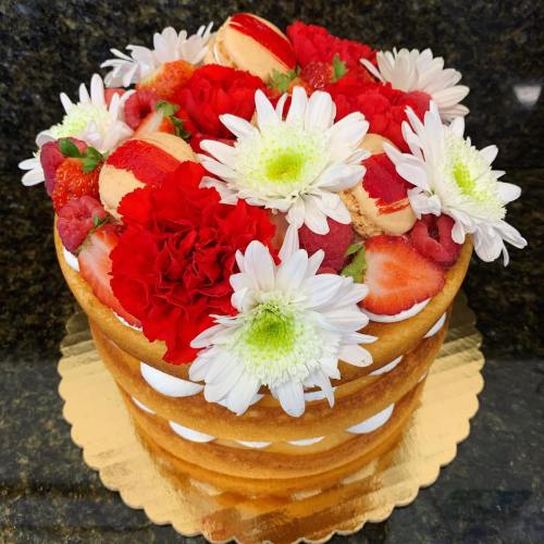 A beautifully decorated cake topped with red flowers, strawberries, raspberries, and macarons on a gold base.