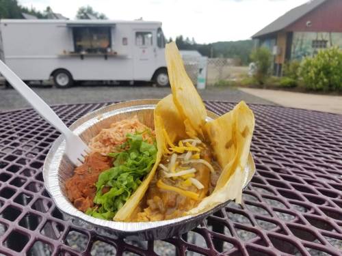A close-up of a meal featuring two tacos, rice, and beans, served in a foil container on a picnic table.