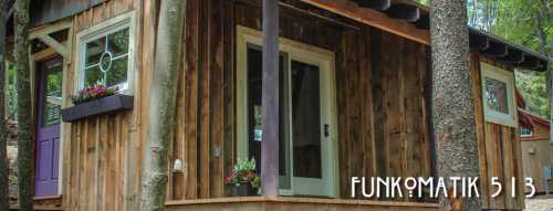 A rustic wooden cabin with large windows and a welcoming door, surrounded by trees and greenery.