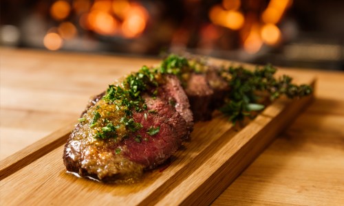 A beautifully cooked slice of beef topped with herbs and sauce, served on a wooden platter with a blurred background.