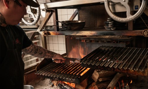A chef using tongs to grill food over an open flame in a commercial kitchen.
