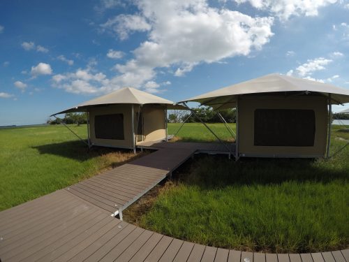 Two canvas tents on a wooden walkway in a grassy field under a blue sky with scattered clouds.