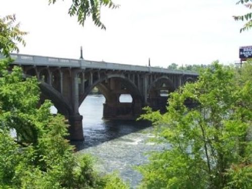 A stone bridge with arches spans a river, surrounded by lush green trees and a clear blue sky.