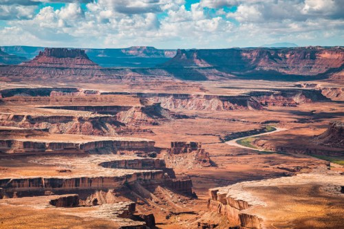 A vast desert landscape with rugged cliffs, canyons, and a winding river under a blue sky with scattered clouds.