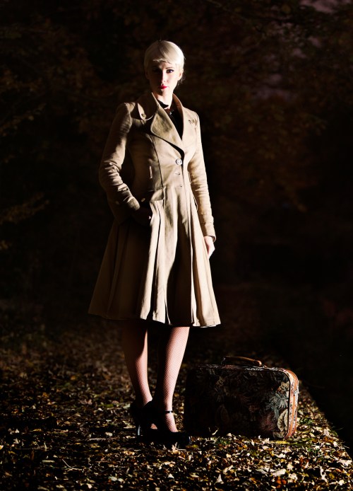 A woman in a beige coat stands on a leaf-covered path, next to a vintage suitcase, with a dark, moody background.