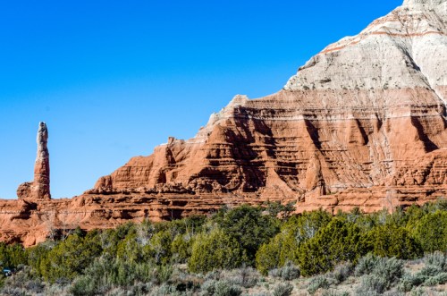 A striking rock formation with a tall spire, surrounded by layered red and white cliffs under a clear blue sky.