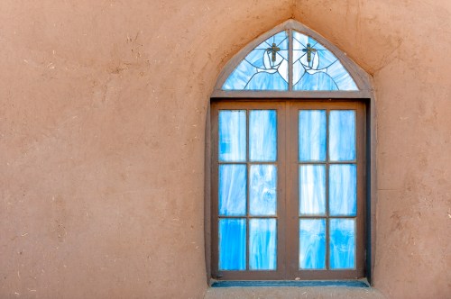 A rustic arched window with blue stained glass set in a textured brown wall.