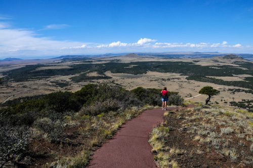 A person walks along a trail overlooking a vast, open landscape with mountains and blue skies in the distance.