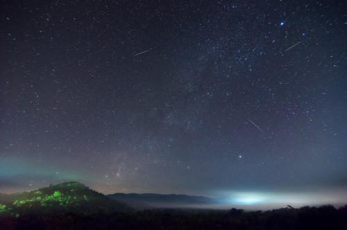 A starry night sky filled with stars and meteors over a misty landscape and distant mountains.