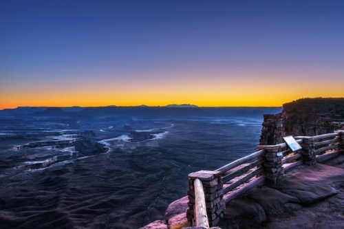 A stunning sunrise over a vast canyon, with a wooden railing in the foreground and mountains in the distance.