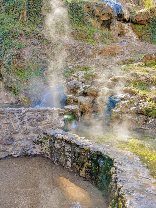 Steam rises from a natural hot spring surrounded by rocky terrain and lush greenery.