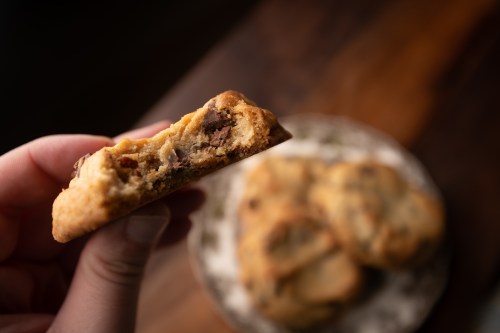 A hand holding a half-eaten cookie, revealing chocolate chips, with more cookies on a plate in the background.