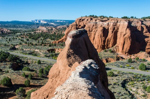 A rocky outcrop rises above a vast desert landscape, with distant cliffs and clear blue skies in the background.
