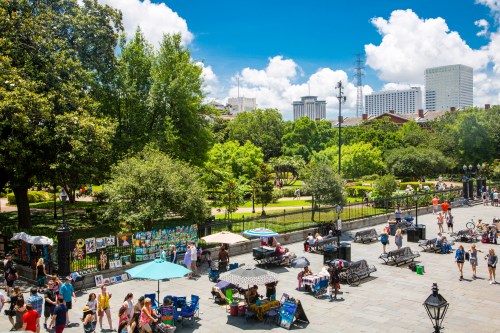 A vibrant park scene with people walking, art vendors, and lush greenery under a bright blue sky.