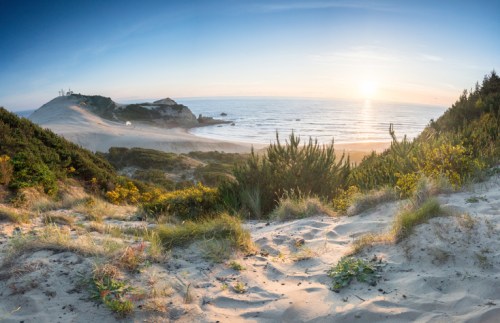 A panoramic view of a sandy beach at sunset, with lush greenery and a distant lighthouse on a coastal hill.