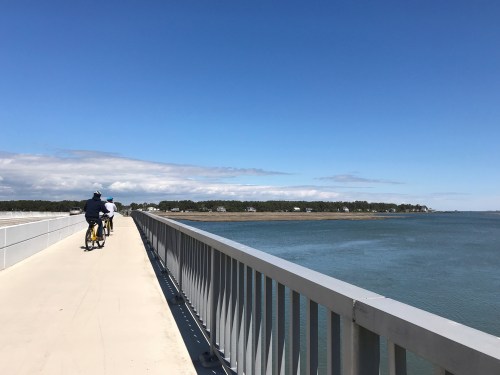 A person rides a yellow bicycle on a bridge overlooking a calm body of water under a clear blue sky.