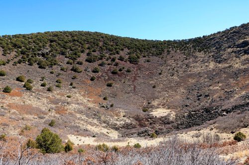 A wide view of a volcanic crater with sparse vegetation and rocky terrain under a clear blue sky.