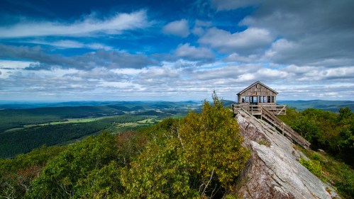 A wooden cabin perched on a rocky outcrop, surrounded by lush green hills and a dramatic sky with scattered clouds.