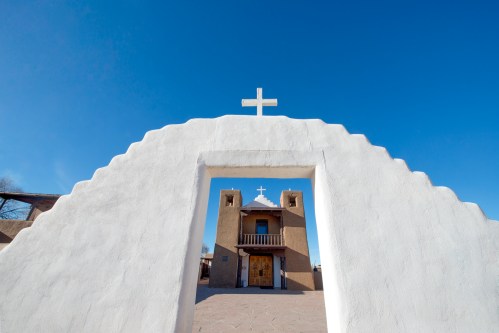 A white archway frames a church with a cross on top, set against a clear blue sky.