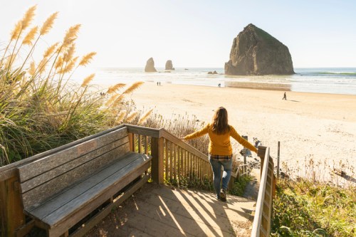 A person in a yellow sweater walks down steps toward a beach with large rock formations and people in the distance.