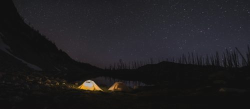 Two tents illuminated by soft light beside a calm lake under a starry night sky.