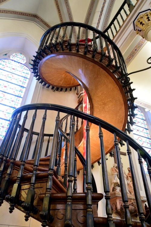 A spiral wooden staircase with ornate black railings, surrounded by stained glass windows.
