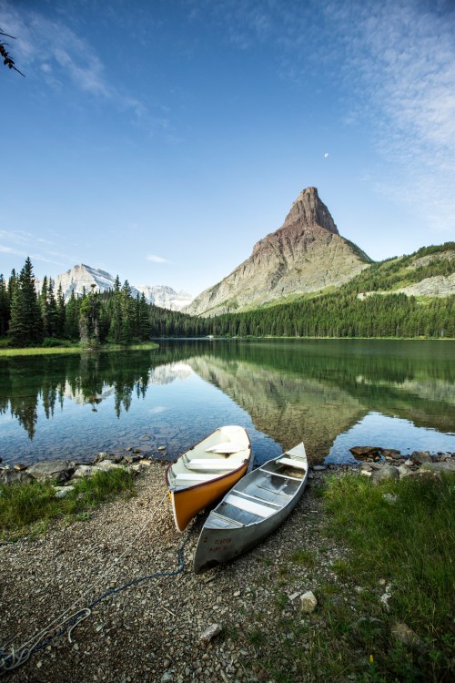 Two canoes on a serene lake, surrounded by lush trees and mountains, reflecting in the calm water under a clear sky.