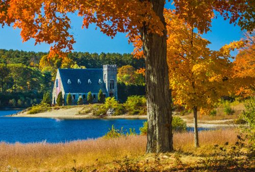 A picturesque scene of a stone house by a lake, surrounded by vibrant autumn foliage and clear blue skies.