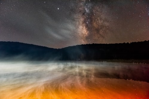 A starry night sky reflects on a misty lake, with vibrant colors and a silhouette of trees in the background.