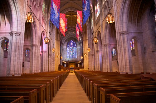 Interior of a grand church with wooden pews, colorful stained glass windows, and vibrant banners hanging from the ceiling.