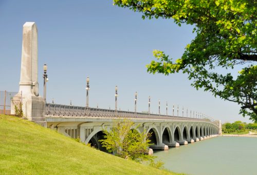 A scenic view of a bridge with arches, lined with lampposts, beside a calm body of water and green grass.