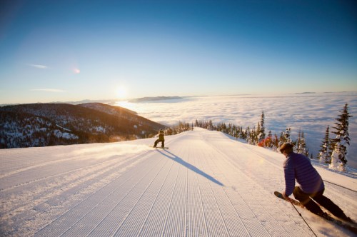 Two skiers carve through freshly groomed snow on a mountain slope, with a stunning sunrise and clouds below.
