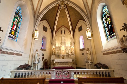 Interior of a church featuring an ornate altar, stained glass windows, and statues, with a high vaulted ceiling.