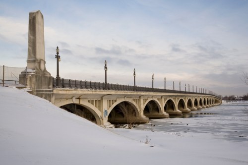 A snow-covered bridge with arches and lampposts spans a frozen river under a cloudy sky.