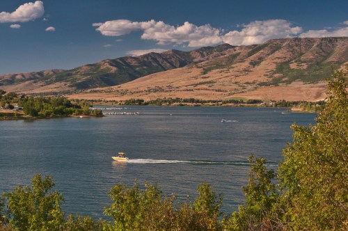 A scenic view of a lake surrounded by mountains, with a yellow boat cruising on the water under a blue sky.