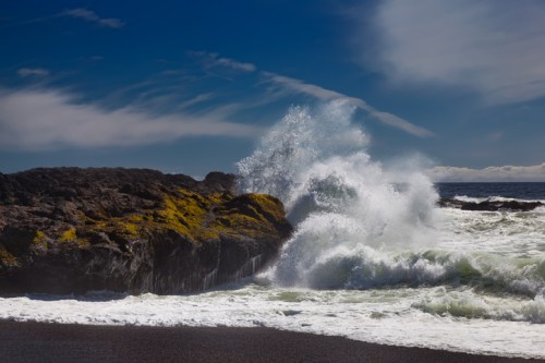 Waves crashing against rocky shore with moss, under a bright blue sky and scattered clouds.