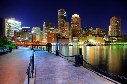 Nighttime view of a city skyline reflecting on water, with illuminated buildings and a waterfront walkway.