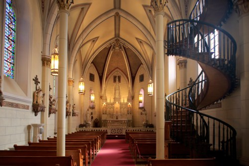 Interior of a church featuring stained glass windows, wooden pews, and a spiral staircase leading to an upper level.
