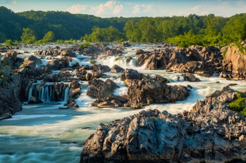 A scenic view of a river flowing over rocky terrain, surrounded by lush greenery and a clear blue sky.
