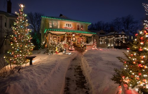 A beautifully decorated house with colorful lights and Christmas trees, surrounded by snow at night.