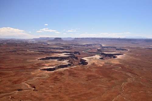 A vast desert landscape with winding canyons and a river, under a clear blue sky with scattered clouds.