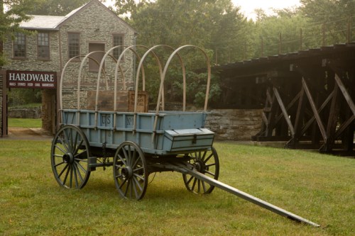 A blue wooden wagon with arches stands on grass near a stone building and a wooden bridge in a historical setting.