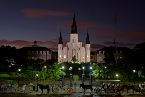 A historic building illuminated at night, with horse-drawn carriages lined up in the foreground and palm trees nearby.