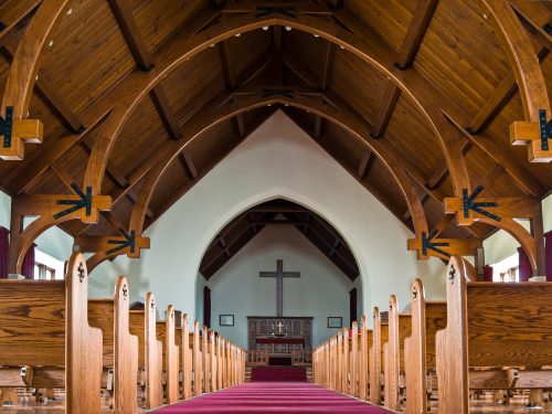 Interior of a church featuring wooden beams, pews, and a large cross at the front, with a red carpet down the aisle.