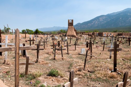 A cemetery with wooden crosses, an old stone tower in the background, and mountains under a clear blue sky.