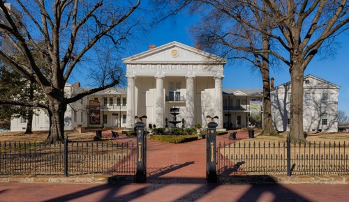 A grand white building with columns, surrounded by trees and a brick pathway, under a clear blue sky.
