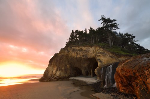 A scenic beach at sunset featuring a rocky cliff, a waterfall, and trees silhouetted against a colorful sky.