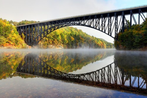 A black steel bridge arches over a misty river, surrounded by colorful autumn foliage reflecting in the water.